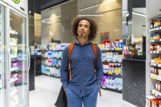 Young man with afro hair stands in a supermarket aisle, thoughtfully comparing products and
