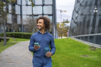 Smiling young Mixed-race man with curly afro hair walking on a path in a modern city park, holding