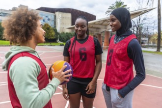 Young diverse athletes wearing sports bibs, holding a basketball, and talking on an urban court,