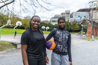 Two diverse young women, one wearing a hijab, smiling and holding a basketball while standing on an