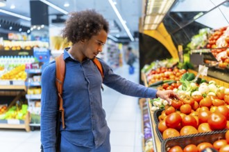 Young man wearing a blue shirt and backpack selecting fresh red tomatoes from the produce aisle,