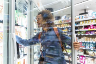 Young black man browsing frozen food in a supermarket aisle, opening a freezer door and selecting