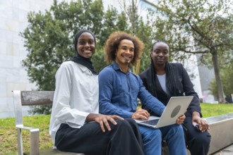 Three diverse students are smiling directly at the camera while sitting outside on a bench. Working