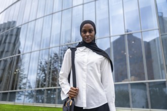 Confident young black woman wearing a hijab and carrying a shoulder bag, standing in front of a