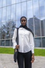 Young black muslim woman wearing a hijab and white shirt, holding a bag and standing confidently
