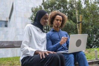 Diverse young man and woman collaborating on a laptop while seated on a park bench, focused on work