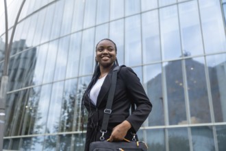 Young black businesswoman smiling and holding a laptop bag, walking with confidence outside a