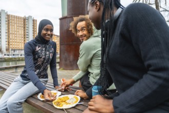 Diverse group of young adults sharing a casual outdoor meal on a city park bench, smiling, talking