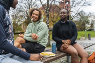 Group of diverse young adults sitting on a park bench, sharing a meal and enjoying casual