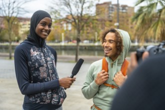 Female journalist wearing a hijab holding a microphone, interviewing a cheerful young man with