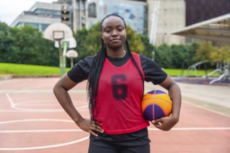 Confident young black woman athlete with braids wearing a red jersey, holding a basketball, and