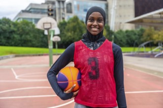 Young muslim woman wearing a hijab and sports uniform, holding a basketball and smiling confidently