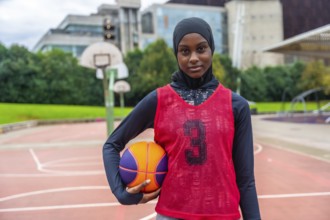 Young muslim woman wearing a hijab, long sleeve shirt, and basketball bib, holding a basketball