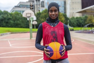 Young woman in a hijab and sports vest standing on an outdoor basketball court, holding a colorful