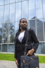 Confident black businesswoman standing outdoors, holding a briefcase, looking away while waiting,