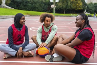 Diverse young adult friends wearing athletic jerseys sitting on an urban outdoor basketball court,