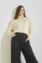 A woman leaning on bright wall in a trendy outfit consisting of a light sweater and dark wide-leg