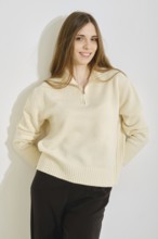A young woman poses in a stylish, cream-colored sweater paired with black pants. She looks with