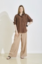 A woman in a trendy and practical outfit, featuring a brown shirt and loose cream trousers stands