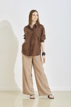 A woman in a trendy brown shirt and beige wide-leg pants posing over white background. Perfect for