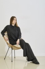 A woman sits gracefully in a wicker chair, wearing a trendy black cardigan and loose pants. The