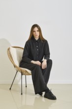 A model sits in a stylish chair, wearing a trendy black shirt and loose pants. Her outfit