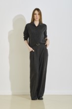 A woman stands in a trendy dark outfit, featuring a button-up top and loose pants. She showcases