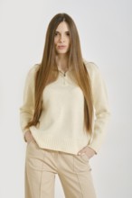 A woman in a cream sweater poses with her hands in pockets of beige pants. Her long hair flows