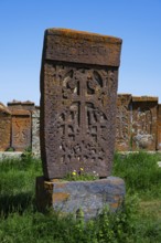 A detailed Armenian crossstone with engravings in front of several others, surrounded by grass