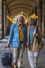 Happy senior couple walking hand in hand through a european arcade, laughing and pulling a suitcase