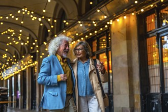 Senior couple walking arm in arm under string lights, happily talking while enjoying an evening out