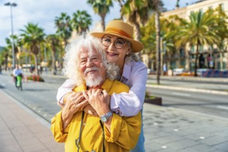 Senior couple embracing and smiling at the camera on a sunny city promenade with palm trees and