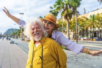 Senior couple laughing as husband gives wife a piggyback through sunlit city streets lined with