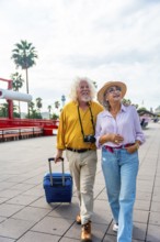 Senior couple walking on a city promenade, smiling and looking up, carrying a suitcase and a