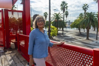 Senior woman standing on red bridge at a sunny harbor, smiling confidently in glasses while