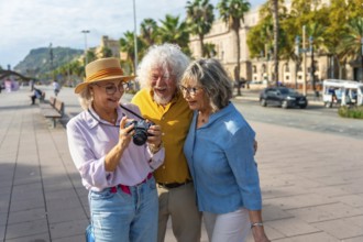 Three happy senior friends are standing on a city street, looking at images on a camera's screen,