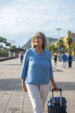 Happy senior woman exploring a city, actively walking with a suitcase, enjoying her vacation and