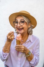 Happy senior woman wearing a straw hat and stylish glasses, smiling while eating a refreshing ice