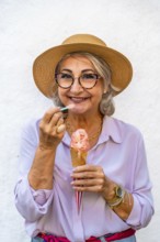 Happy senior woman wearing a straw hat and glasses smiling while eating a refreshing ice cream cone