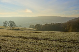 View over a frosty field and distant hills with wind turbines on the horizon at dawn, Seckmauern,