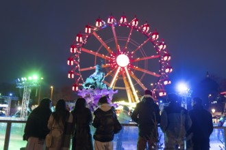 Several people watch ice skaters in front of the Neptune Fountain and a Ferris wheel at the Berlin