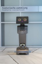 A robot stands in front of a sign for the historical collection on marble floor, robot personality