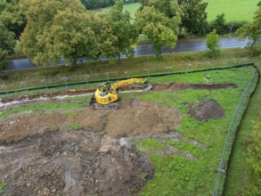 Excavator on a construction site with earthworks next to a road and trees, Bau PV