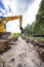 A yellow excavator digs a deep ditch surrounded by nature and a cloudy sky, Bau PV
