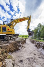 A yellow excavator carries out trenching work on a construction site under a cloudy sky, Bau PV