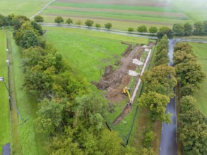 Aerial view of a construction site with excavator, surrounded by fields and trees, construction of