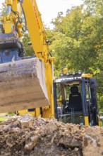 Close-up of an excavator with a raised grab arm at a construction site in front of trees, PV