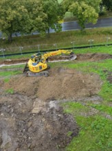 Excavator on a construction site with excavated soil surrounded by greenery and trees, Bau PV