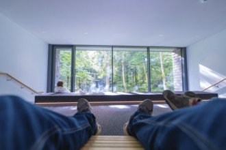 Person relaxing on wooden loungers in a modern room with a large glass front and a view of the