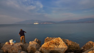 A fisherman fishing on rocky shore with a large sailing ship in the background in grey weather,